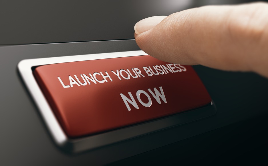 Finger pressing a red button with the text launch your business now. Concept of new venture or startup. Composite image between a hand photography and a 3D background.